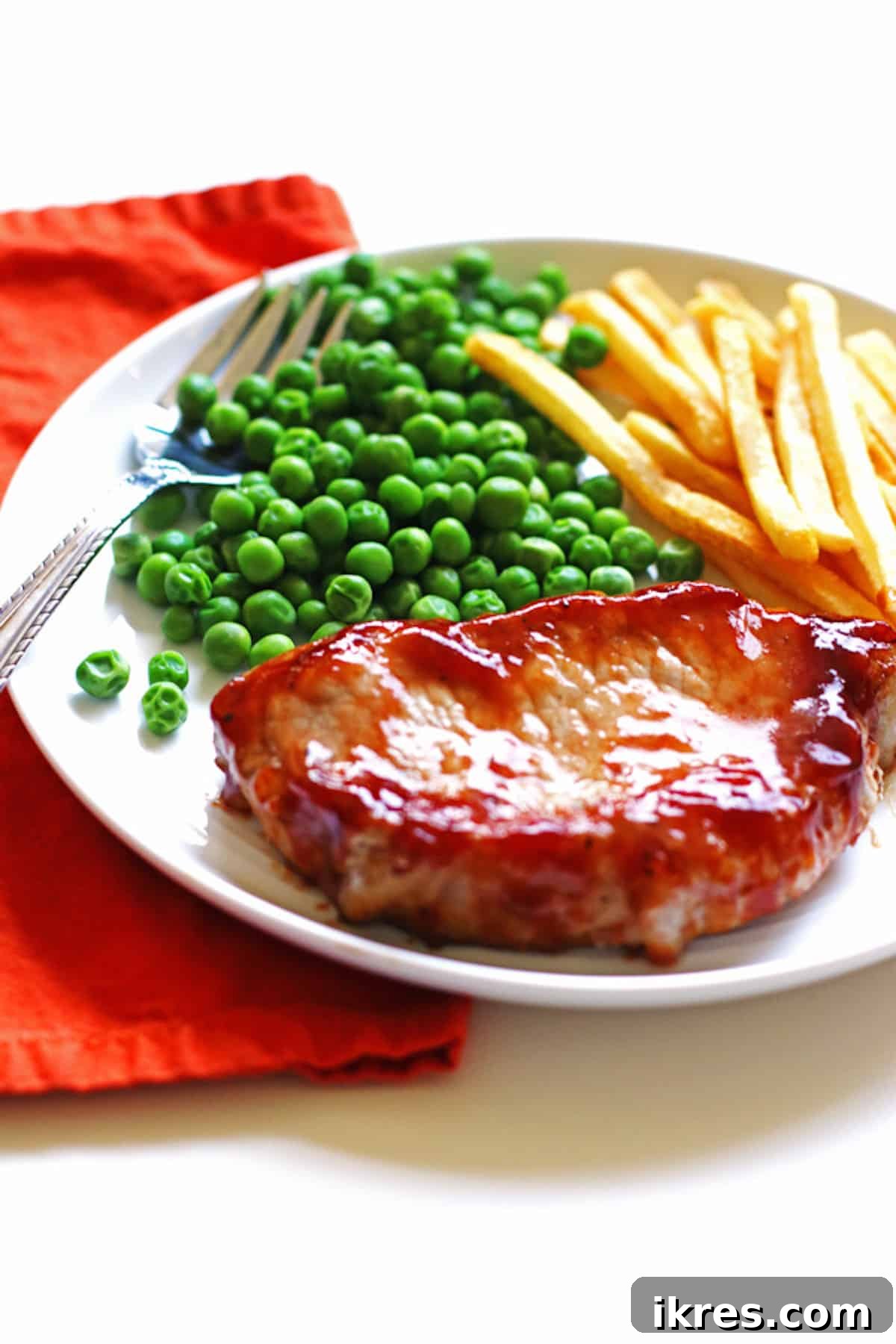 Smoky Grilled Pork Chops 3 A white plate featuring a perfectly cooked barbecue pork chop, accompanied by vibrant green peas and crispy golden french fries.