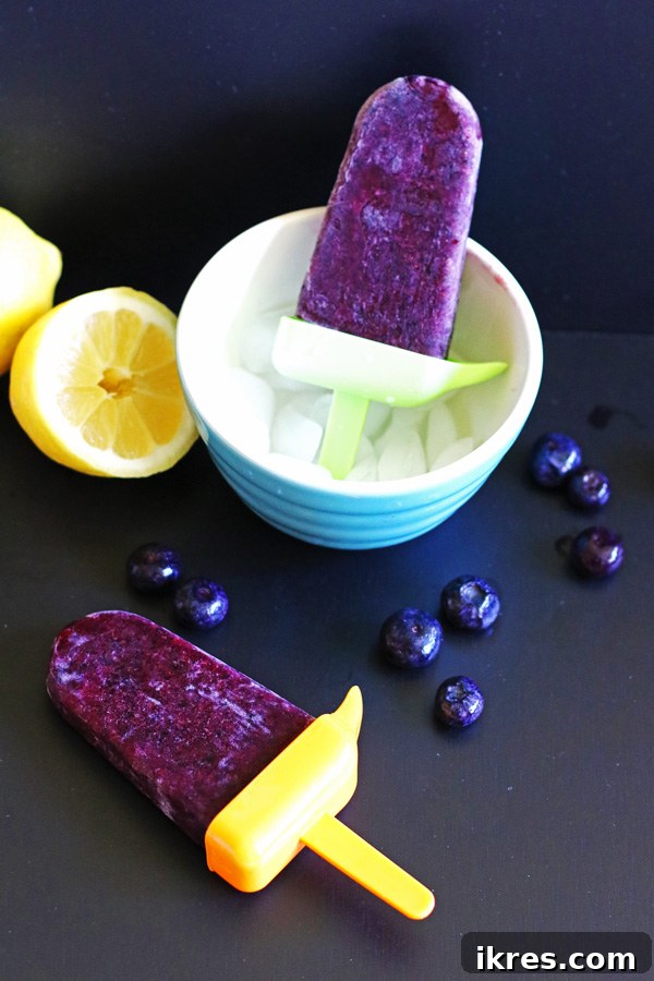 Two homemade blueberry lemon popsicles, one standing in a bowl of ice and another laying on a black surface, ready to be enjoyed.