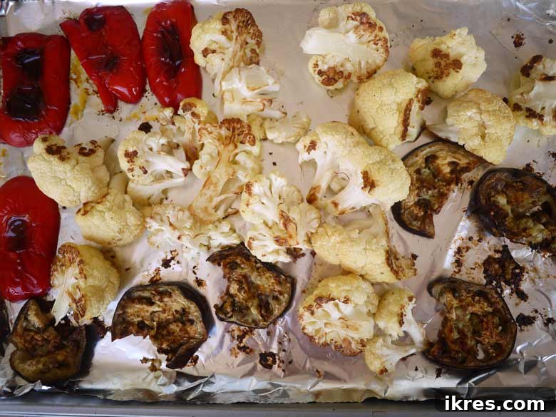 Freshly cut cauliflower florets, sliced red pepper, and eggplant pieces neatly arranged on a baking sheet, glistening with olive oil, ready for roasting.
