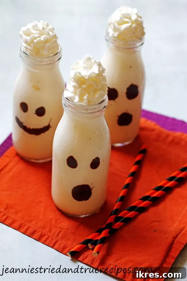Monster Milkshake Creations 2 Two clear glasses showcasing vanilla milkshakes decorated as cute ghosts, set against a festive Halloween backdrop. The ghosts have melting chocolate chip faces and are topped with fluffy white whipped cream.