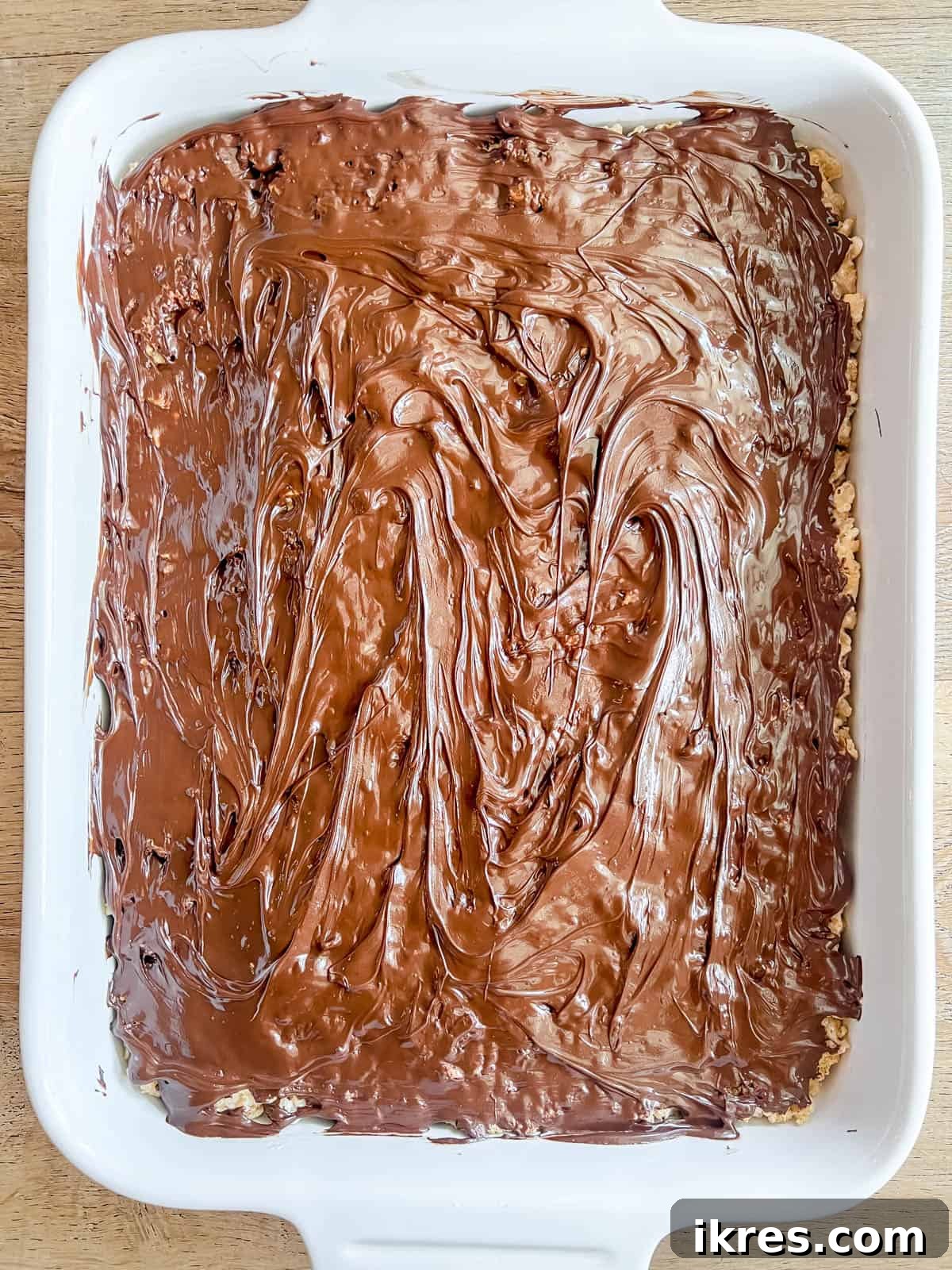 Melted chocolate is being carefully spread over the cooled S'mores Rice Krispie Treat base in the pan, creating a smooth and even layer.