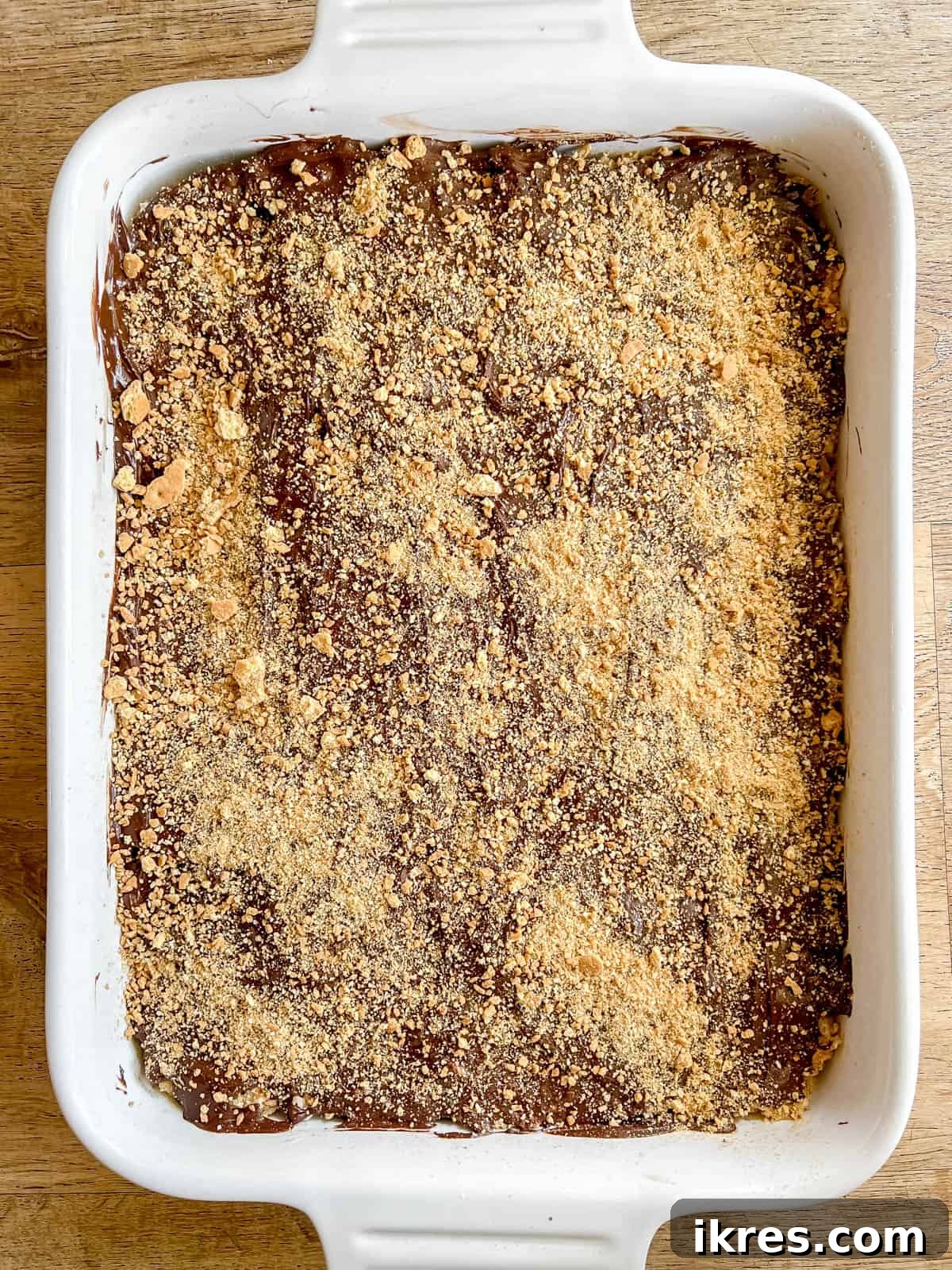 The remaining crushed graham crackers are being sprinkled generously over the freshly spread melted chocolate layer on the S'mores Rice Krispie Treats.