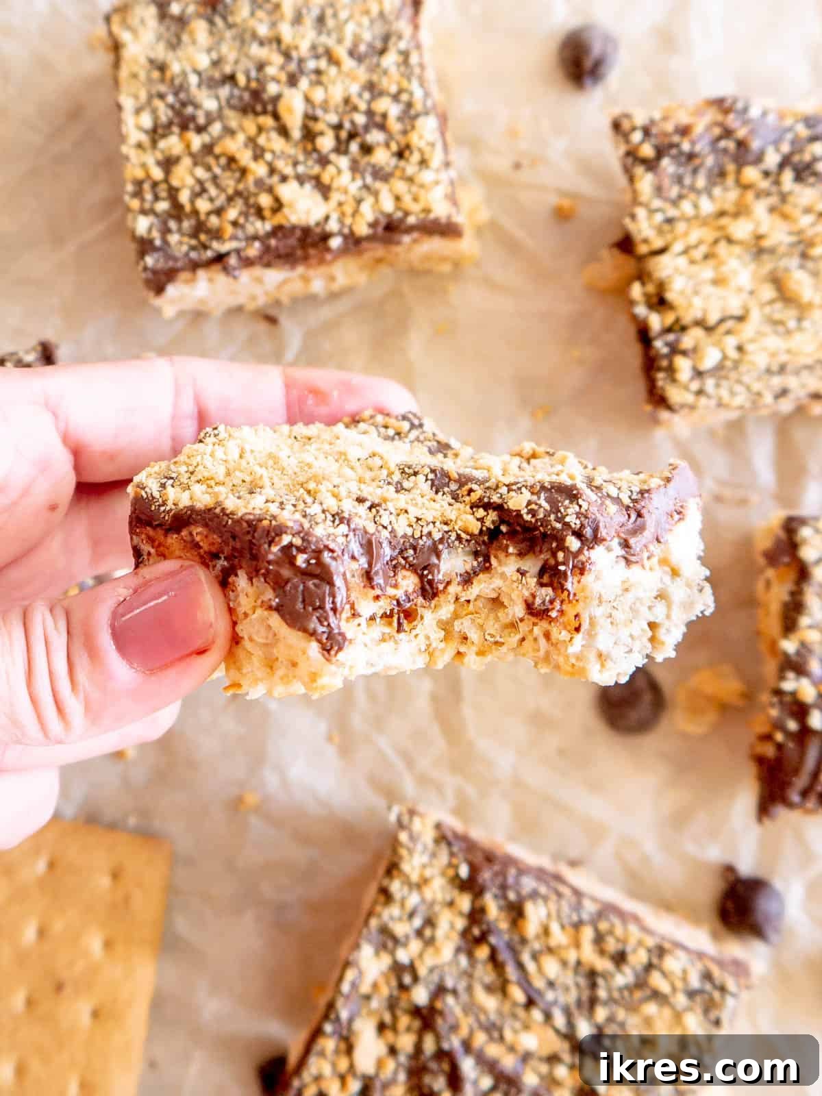 A S'mores Rice Krispie Treat with a bite taken out of it, revealing the crispy cereal, gooey marshmallow, thick chocolate layer, and graham cracker crumbs, emphasizing its delicious texture.