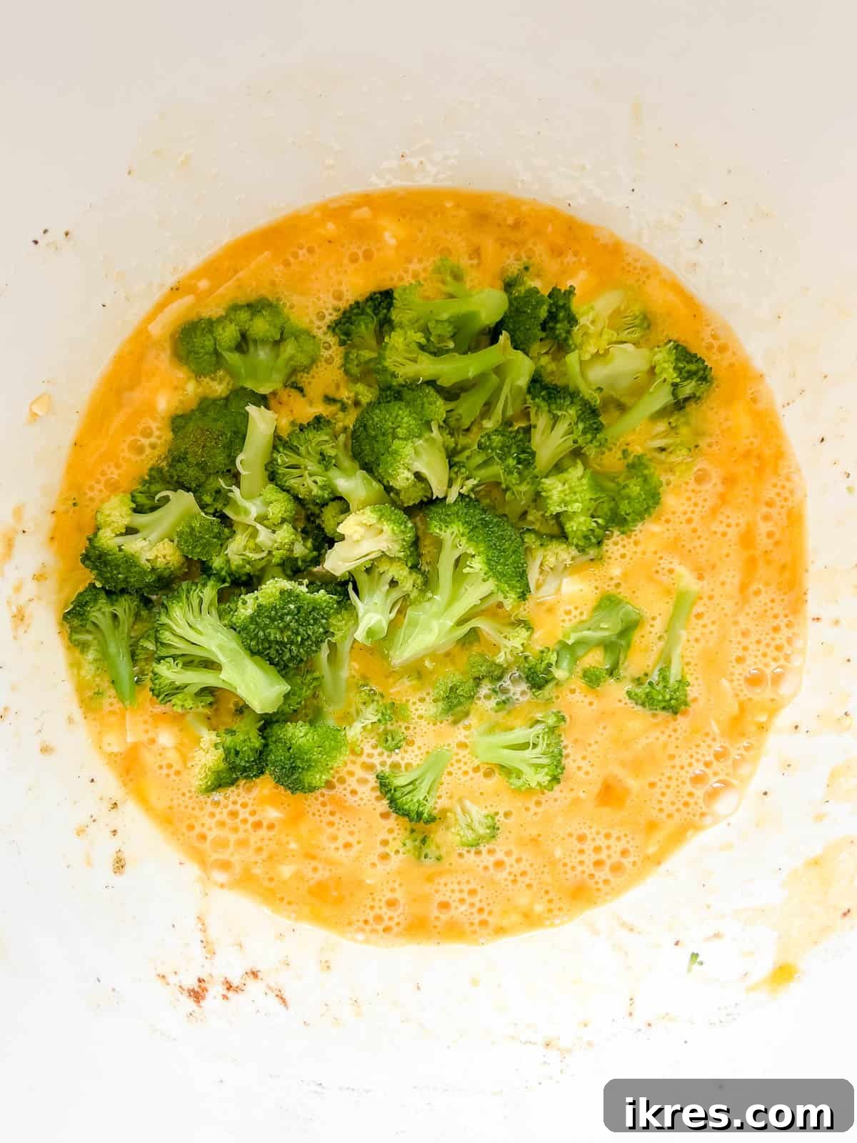 Steamed broccoli florets being folded into the seasoned egg and cheese mixture in a large bowl.
