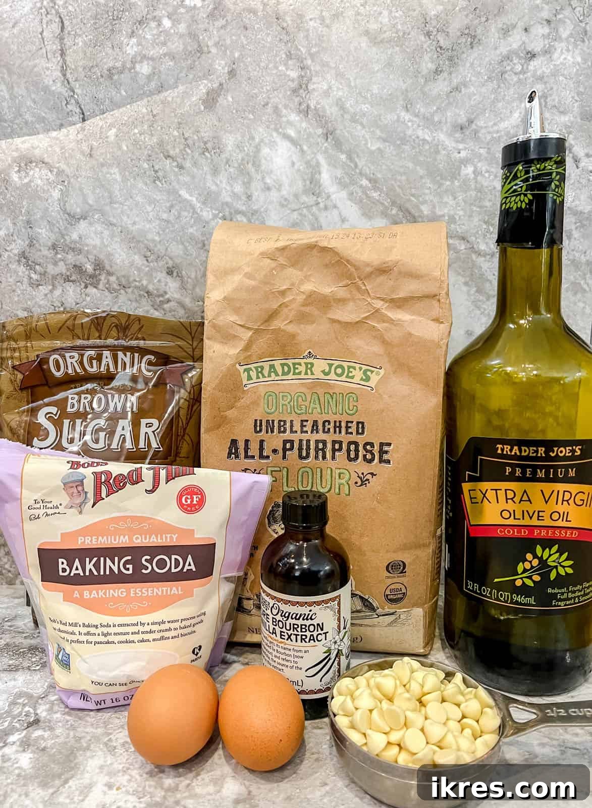 A flat lay photograph showcasing all the fresh ingredients required for making homemade white chocolate chip cookies, including flour, brown sugar, eggs, vanilla, olive oil, and premium white chocolate chips.