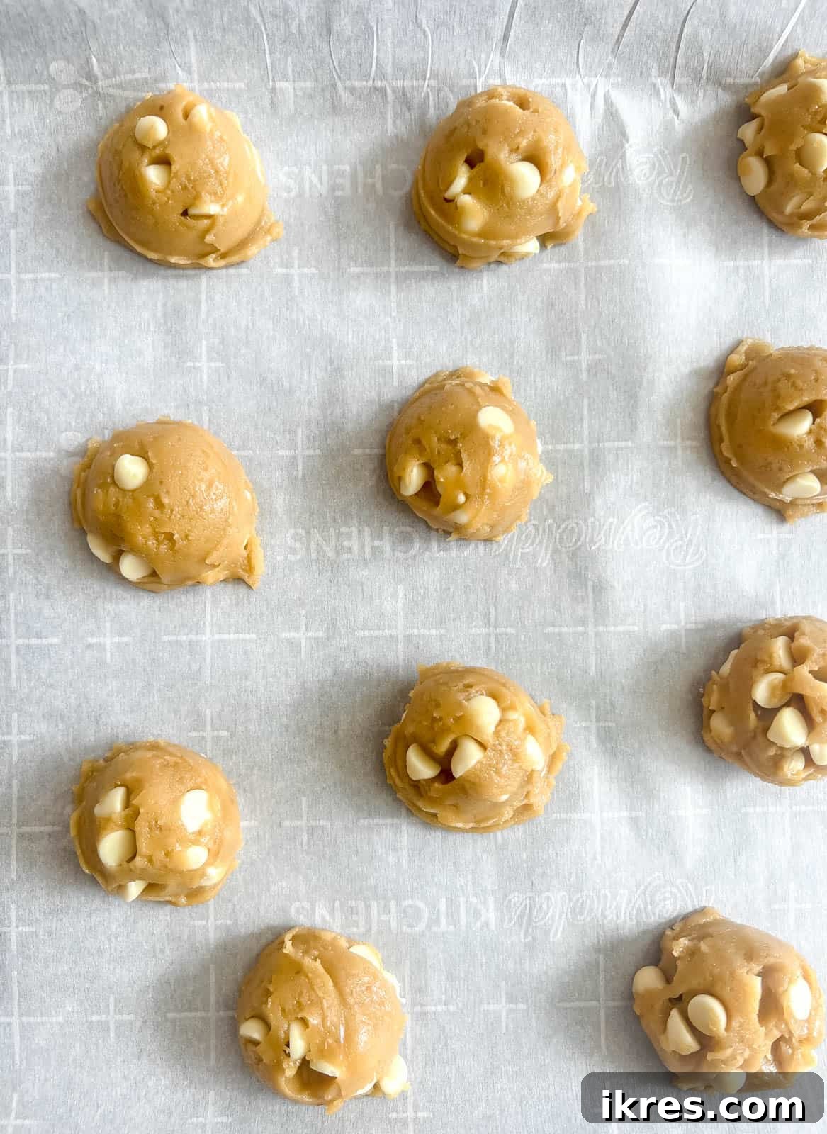 Raw white chocolate chip cookie dough balls, perfectly scooped and placed on a parchment-lined baking sheet, awaiting their transformation in the oven.