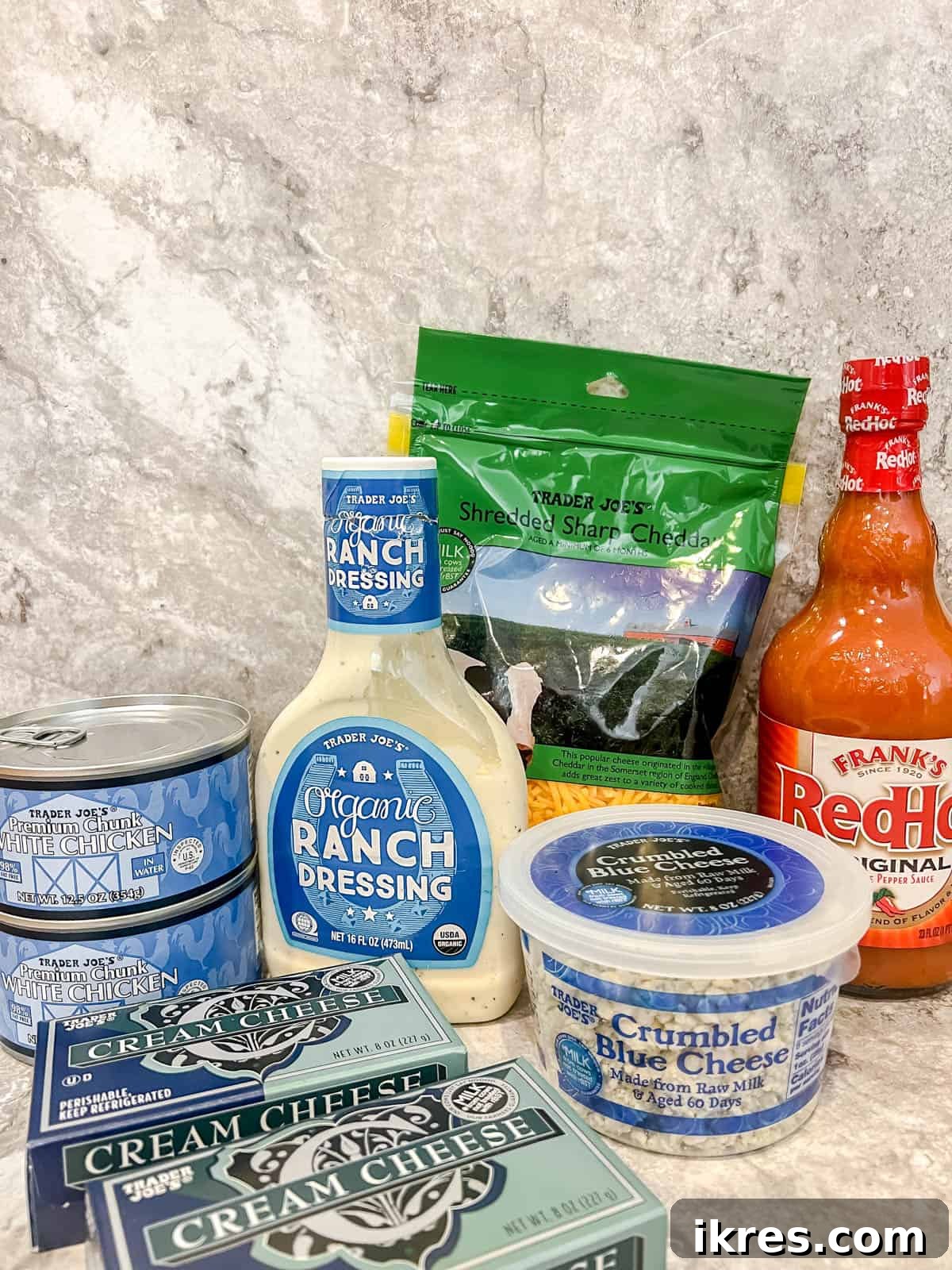 Individual ingredients for buffalo chicken dip including canned chicken, cream cheese, hot sauce, ranch, and blue cheese crumbles laid out on a table.