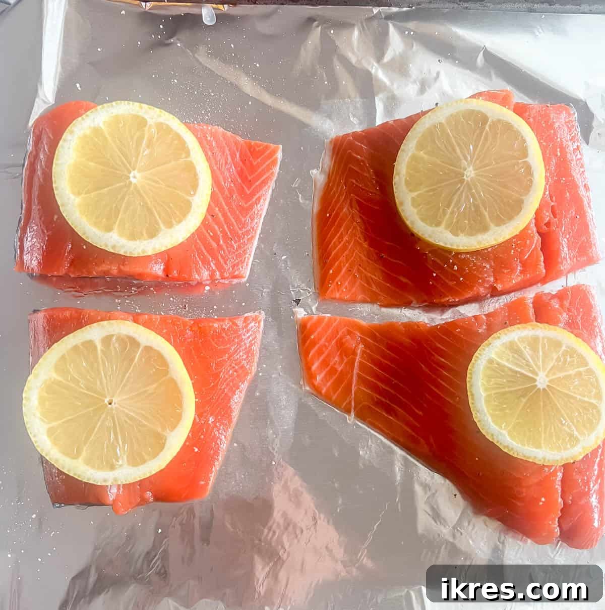 Raw salmon fillets seasoned with salt, pepper, and lemon slices on aluminum foil before baking.