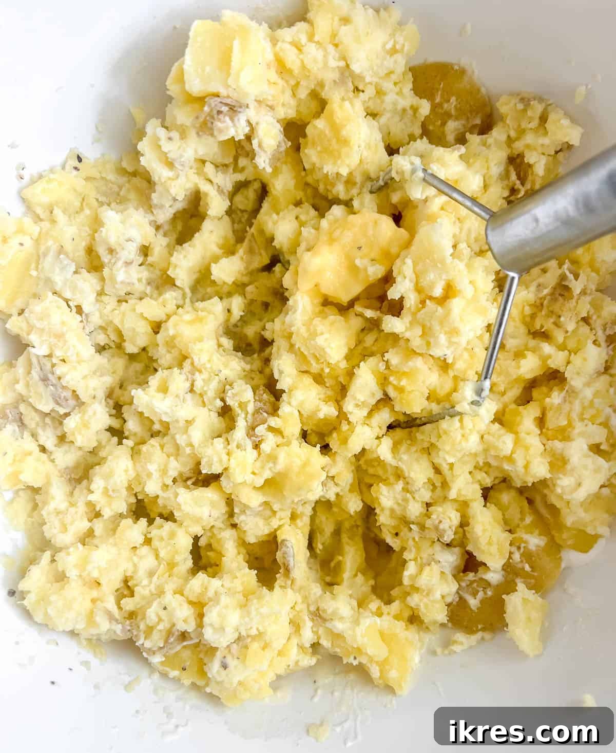 Mashed potatoes being worked with a potato masher in a bowl, showing the creamy texture developing.