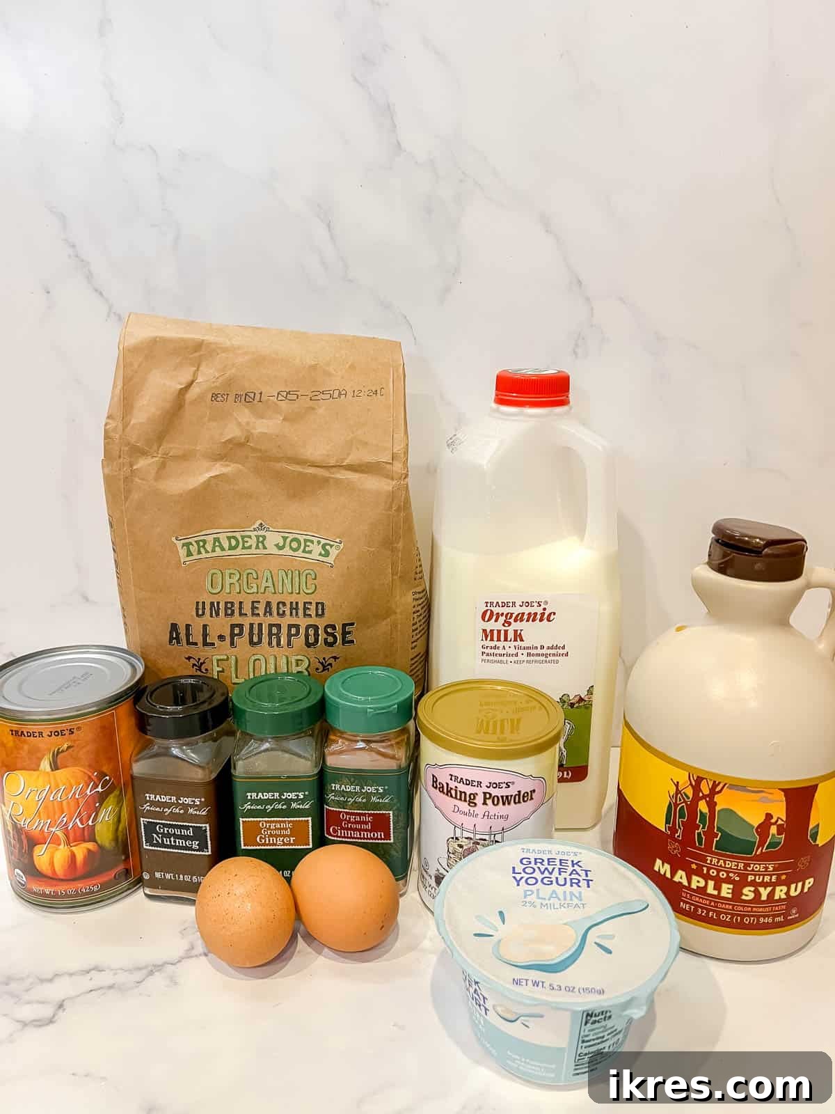 All the necessary ingredients for healthy pumpkin pancakes laid out on a table: eggs, Greek yogurt, pumpkin puree, maple syrup, baking powder, flour, cinnamon, ginger, nutmeg, and milk.