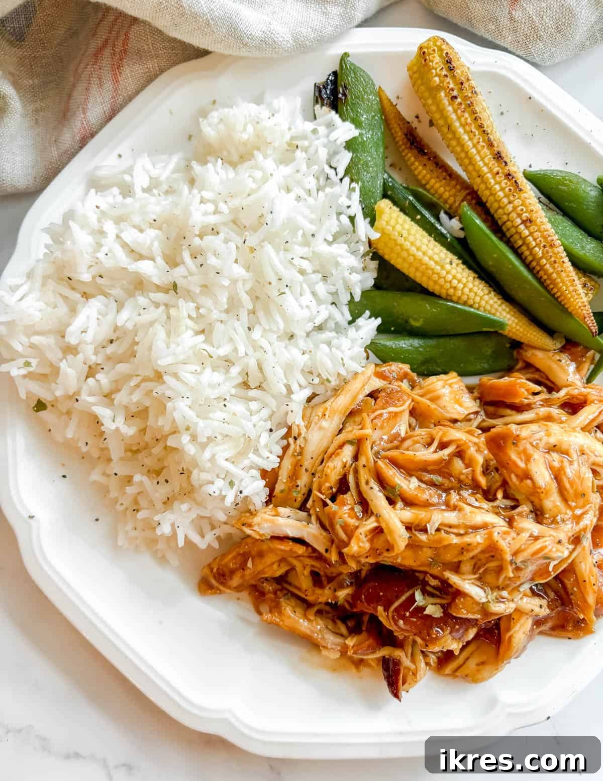 A close-up of a plate with slow cooker shredded honey garlic chicken, garnished with green onions, served alongside rice and a colorful mix of snap peas and corn.