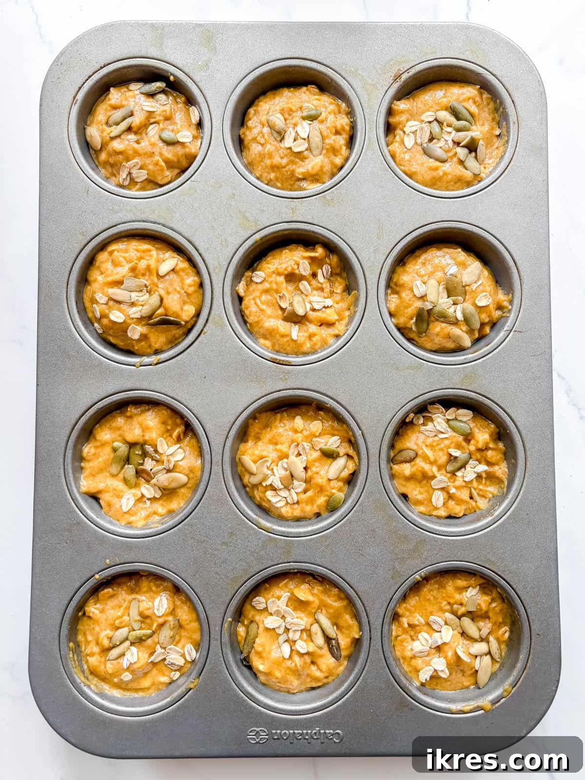 Plant-Based Pumpkin Spice Oat Muffins 7 Pumpkin oatmeal muffin batter scooped into a muffin tin, sprinkled with oats and pumpkin seeds, ready for baking.