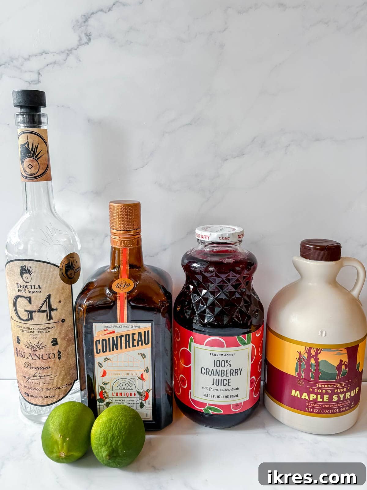 Recipe ingredients laid out on a table, including blanco tequila, Cointreau, cranberry juice, lime juice, and maple syrup.