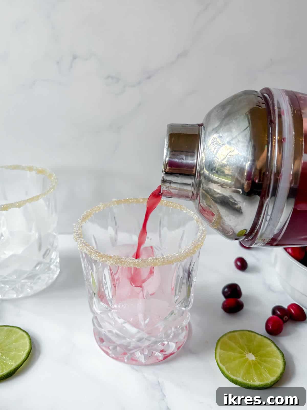 Cranberry margarita mixture being poured from a cocktail shaker into a sugar-rimmed glass filled with ice.