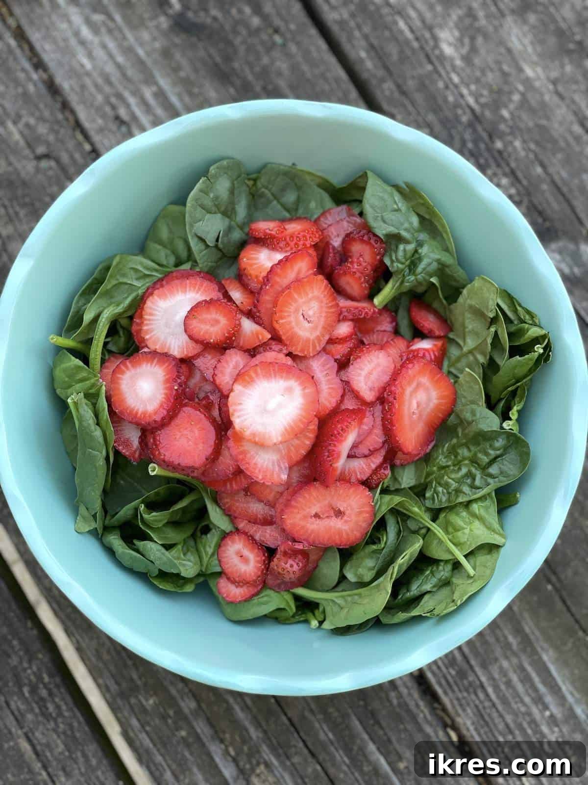 A large serving bowl filled with fresh spinach, topped with vibrant sliced strawberries, ready for further assembly.