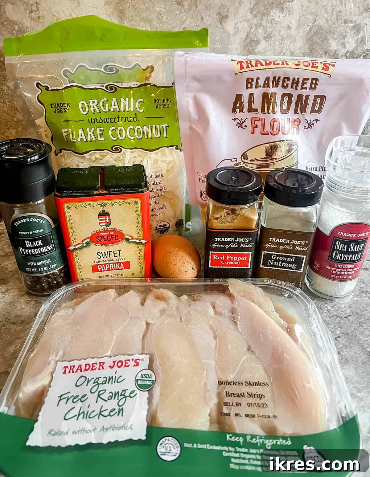 A collection of fresh ingredients laid out on a wooden board, including chicken tenders, almond flour, shredded coconut, egg, and spices, ready for baking paleo chicken tenders.