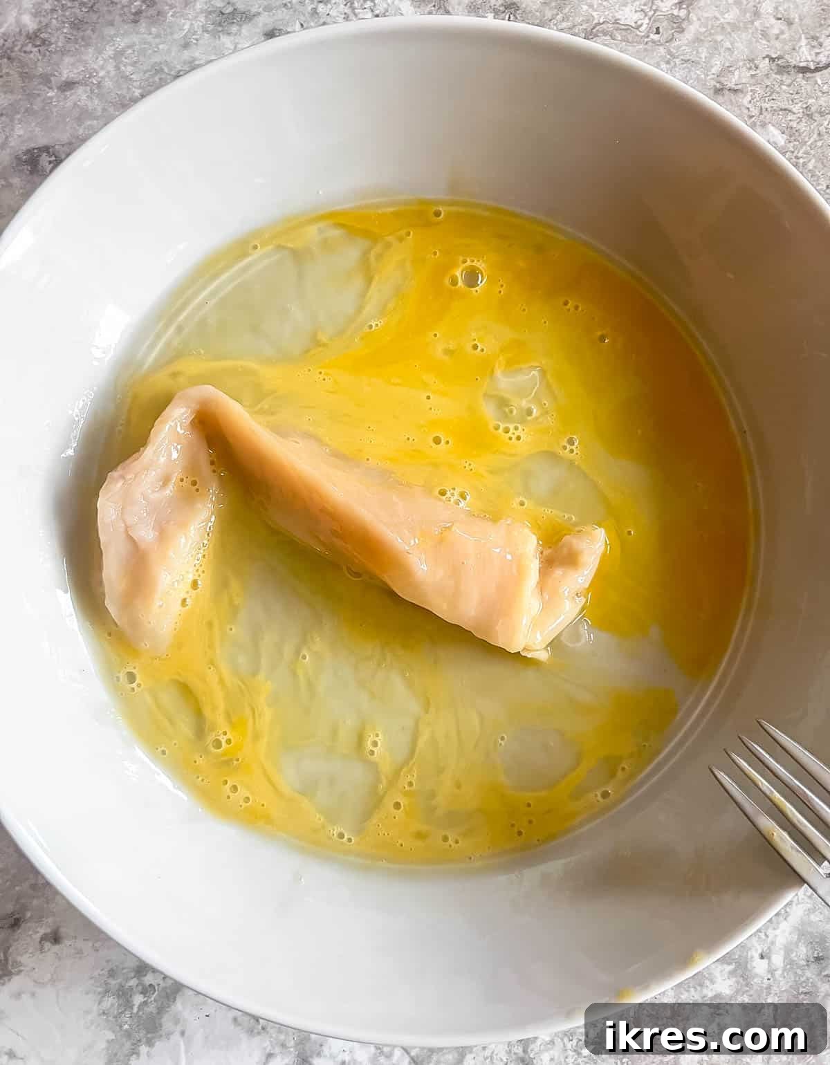 A piece of chicken tender submerged in the whisked egg mixture within a bowl, ensuring a full coating.