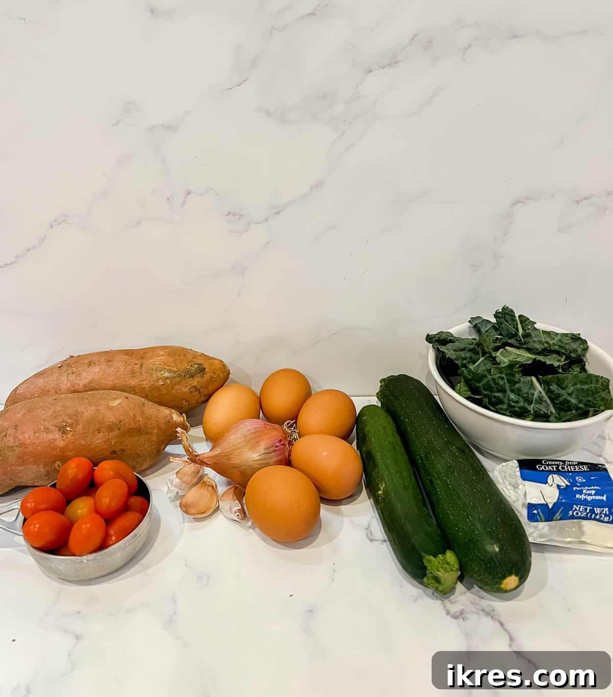 An organized display of all ingredients required to make Zucchini and Kale Sweet Potato Crust Quiche, including eggs, shallots, garlic, zucchini, kale, sweet potatoes, cherry tomatoes, and goat cheese on a wooden board.