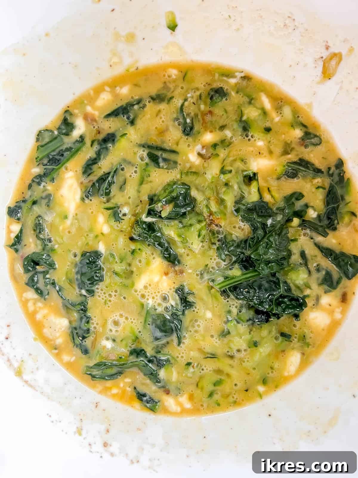 Cooked kale and zucchini being gently mixed into the prepared egg and milk custard mixture, ready to be poured into the crust.