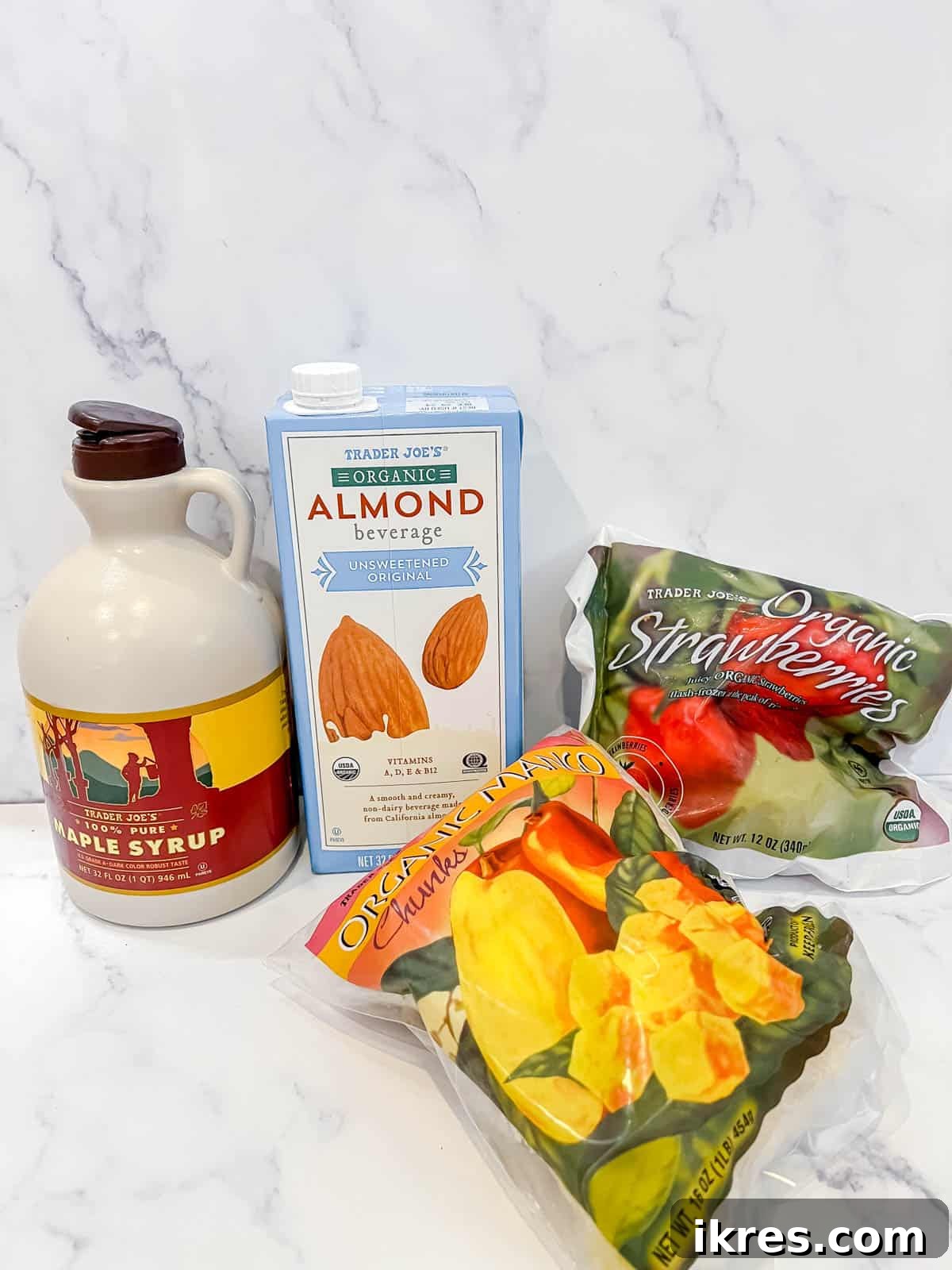 A flat lay showing the simple ingredients needed to make strawberry mango sorbet: frozen strawberries, frozen mango chunks, a bottle of plant-based milk, and a small jar of maple syrup.