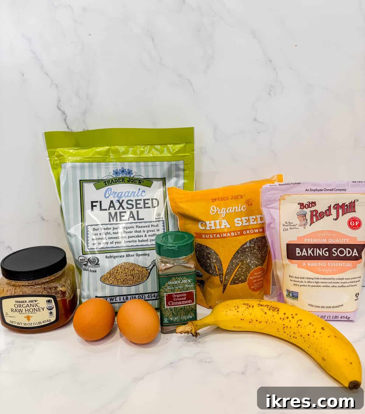 An array of ingredients laid out on a table, including eggs, a banana, ground chia seeds, ground flaxseeds, cinnamon, lemon juice, baking soda, and honey.