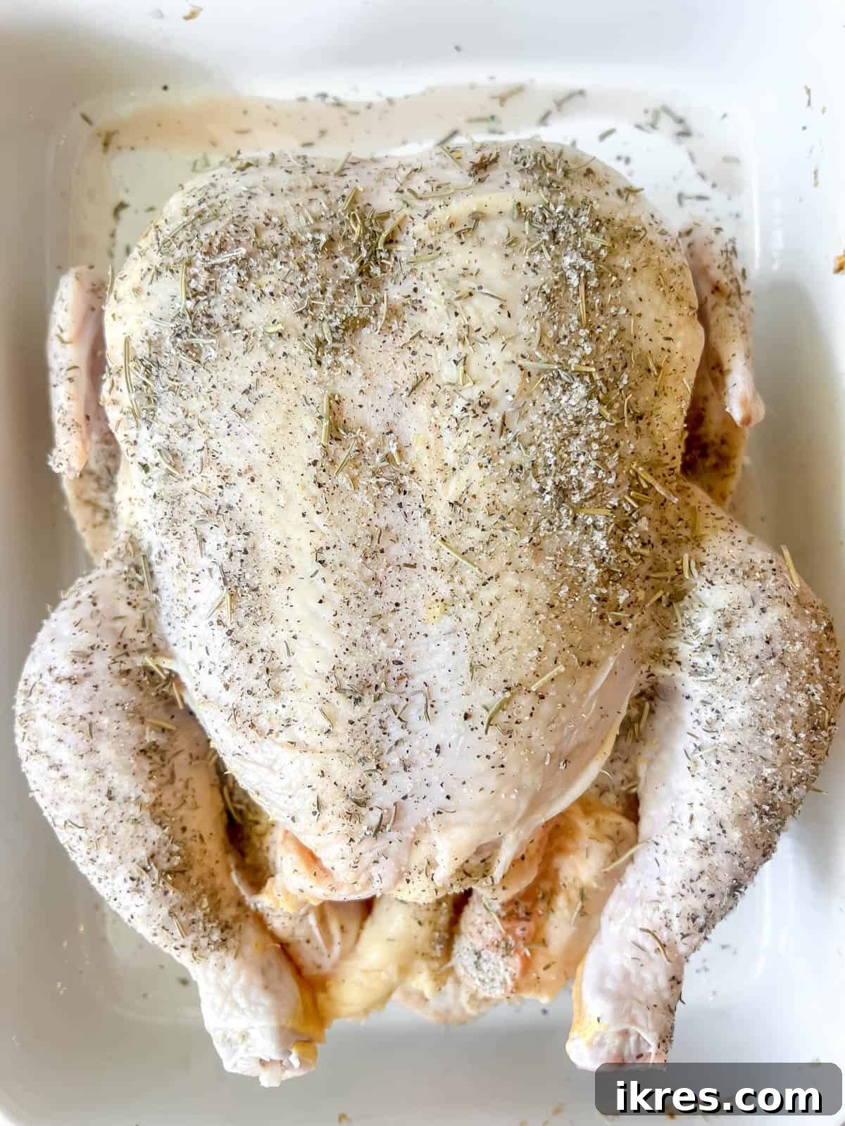 Raw whole chicken in a roasting pan, generously seasoned with the mixed herbs and spices.