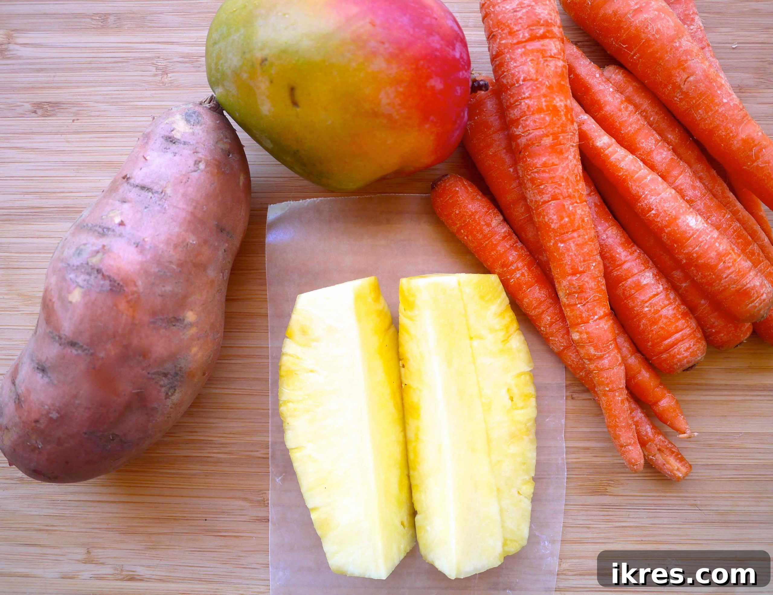 A selection of fresh ingredients laid out: a sweet potato, whole pineapple, several carrots, a ripe mango, and a lemon slice, ready for juicing.