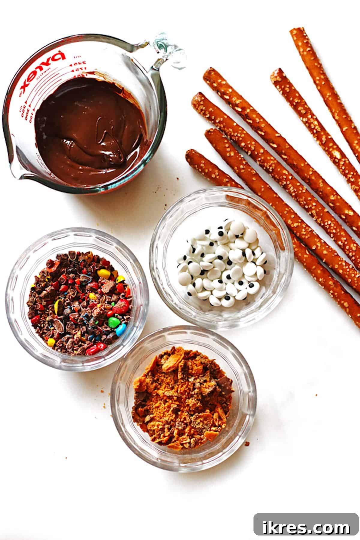 Sweet and Salty Chocolate Pretzel Rods 3 Melted chocolate, pretzel rods, and various crushed candies laid out on a white table, ready for dipping and decorating.