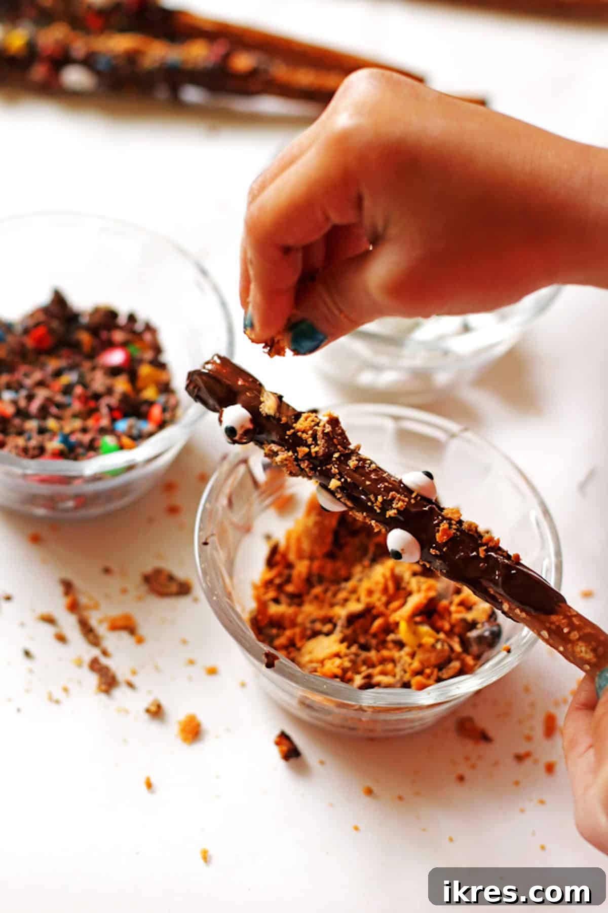 Sweet and Salty Chocolate Pretzel Rods 4 Hands sprinkling colorful crushed candy onto a chocolate-dipped pretzel rod over a white surface.