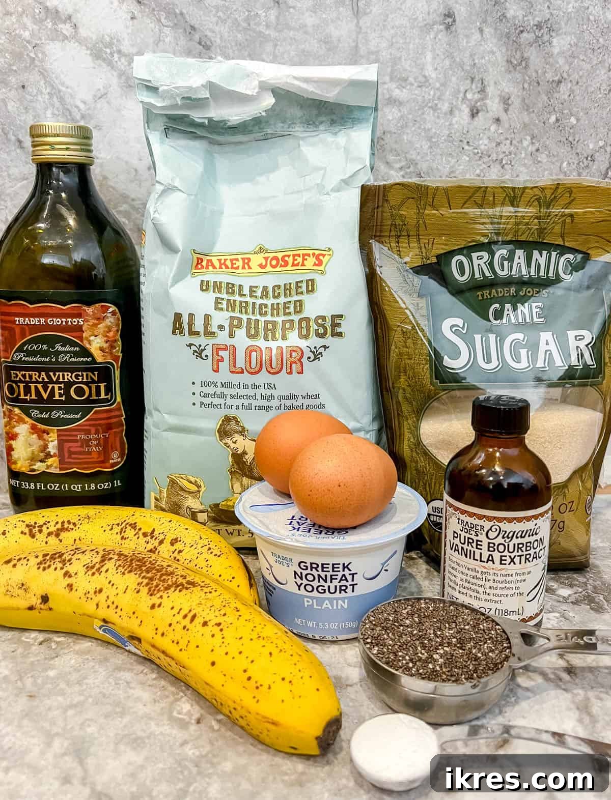 A collection of ingredients laid out, including olive oil, eggs, bananas, Greek yogurt, sugar, flour, baking soda, and chia seeds.