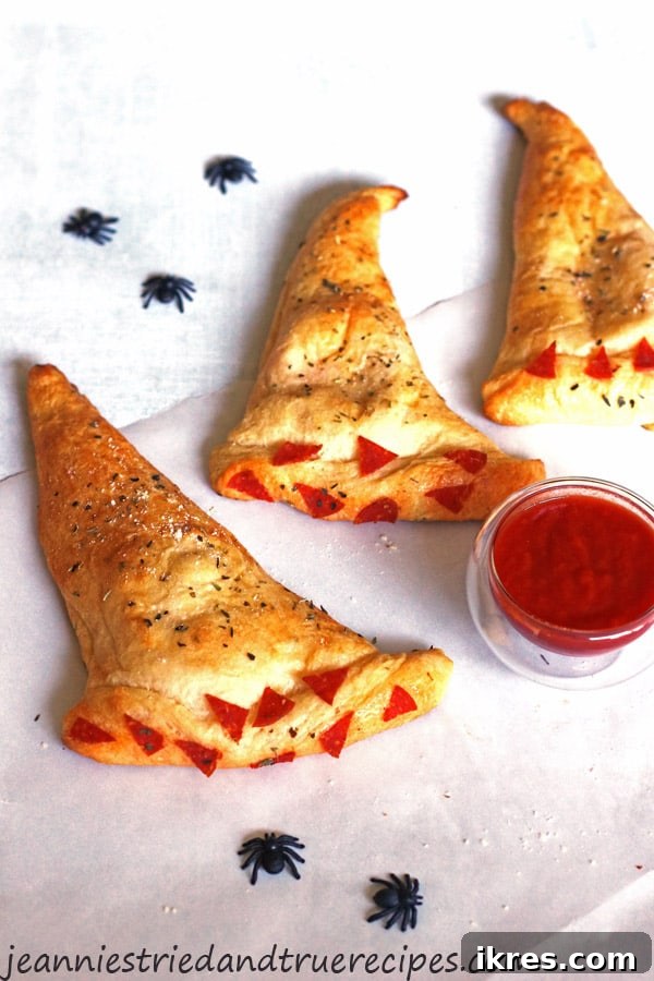 Bewitching Calzone Caps 2 Witch Hat Calzones on a white table with a small bowl of marinara sauce. There are some plastic black spiders on the table for decoration.