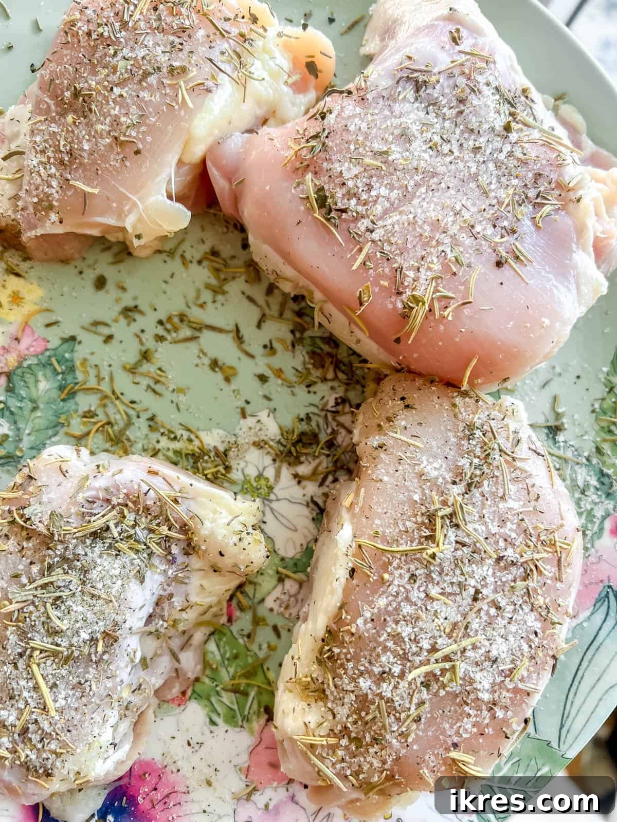 Raw chicken thighs visibly coated with a generous layer of savory herb and spice seasoning, resting before cooking.