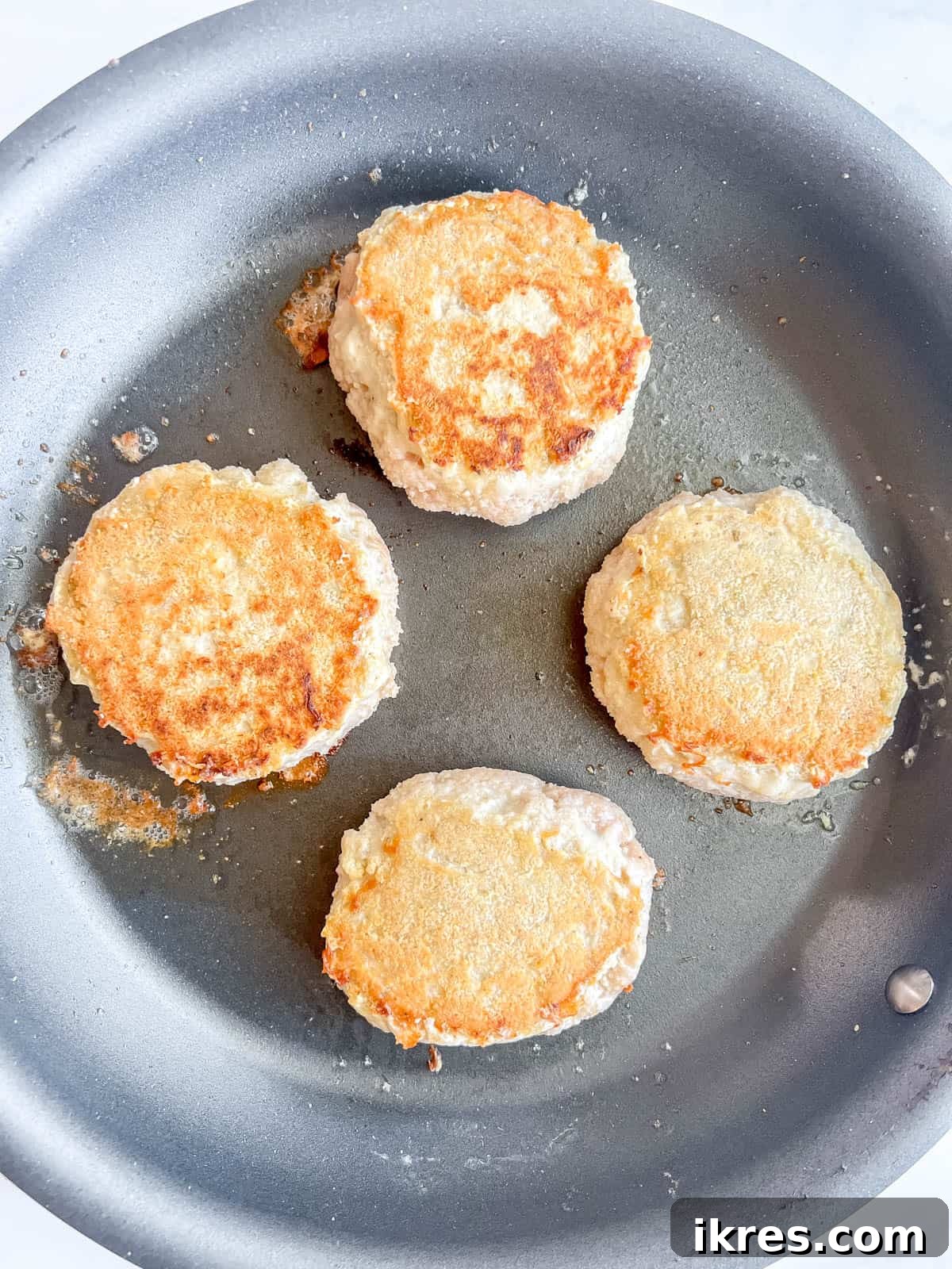 Chicken burgers being flipped in a pan, showing a golden-brown sear on one side.