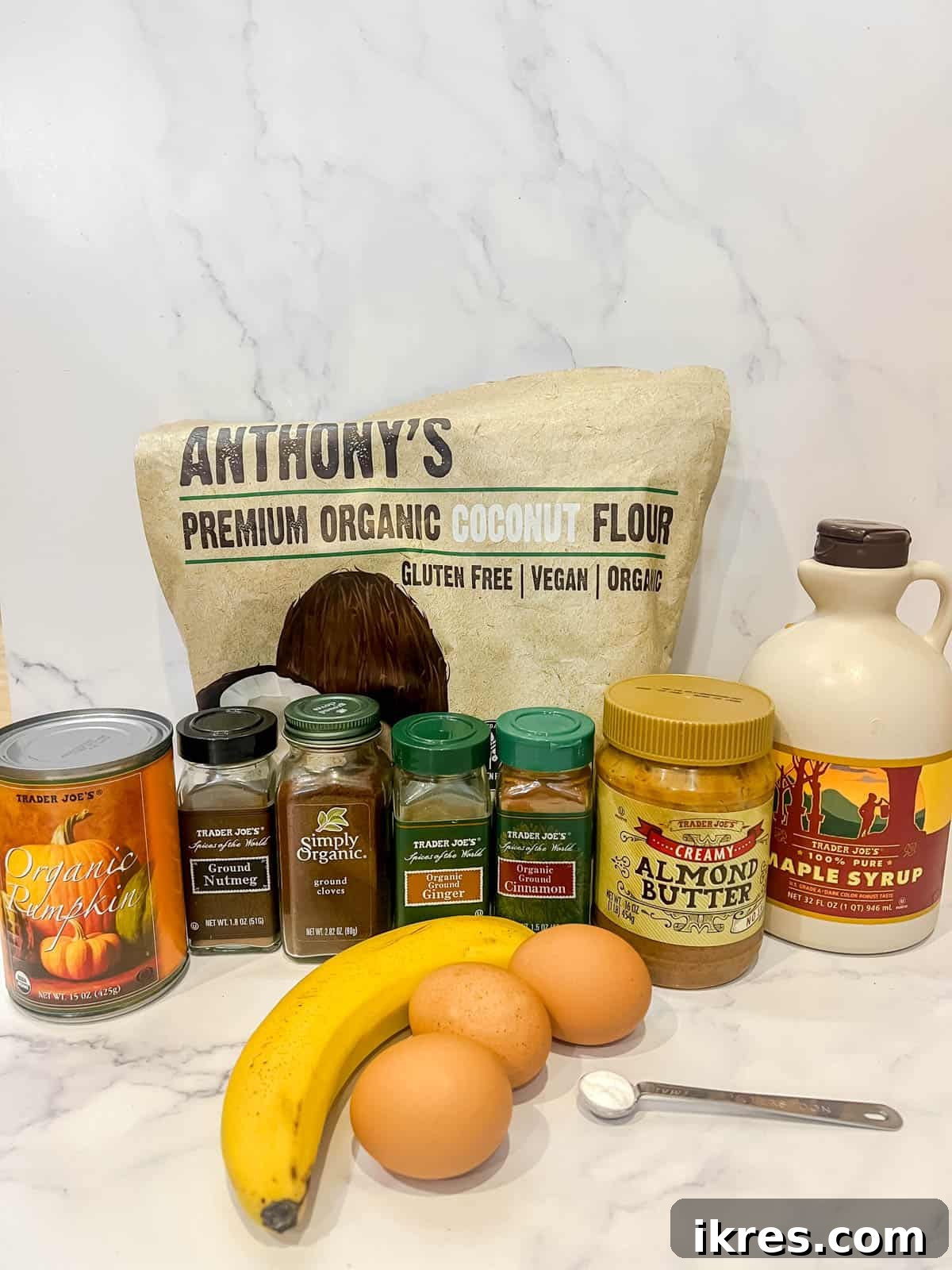 Individual ingredients for Paleo Pumpkin Pancakes laid out: mashed banana, eggs, pumpkin puree, maple syrup, almond butter, coconut flour, and a mix of fall spices.