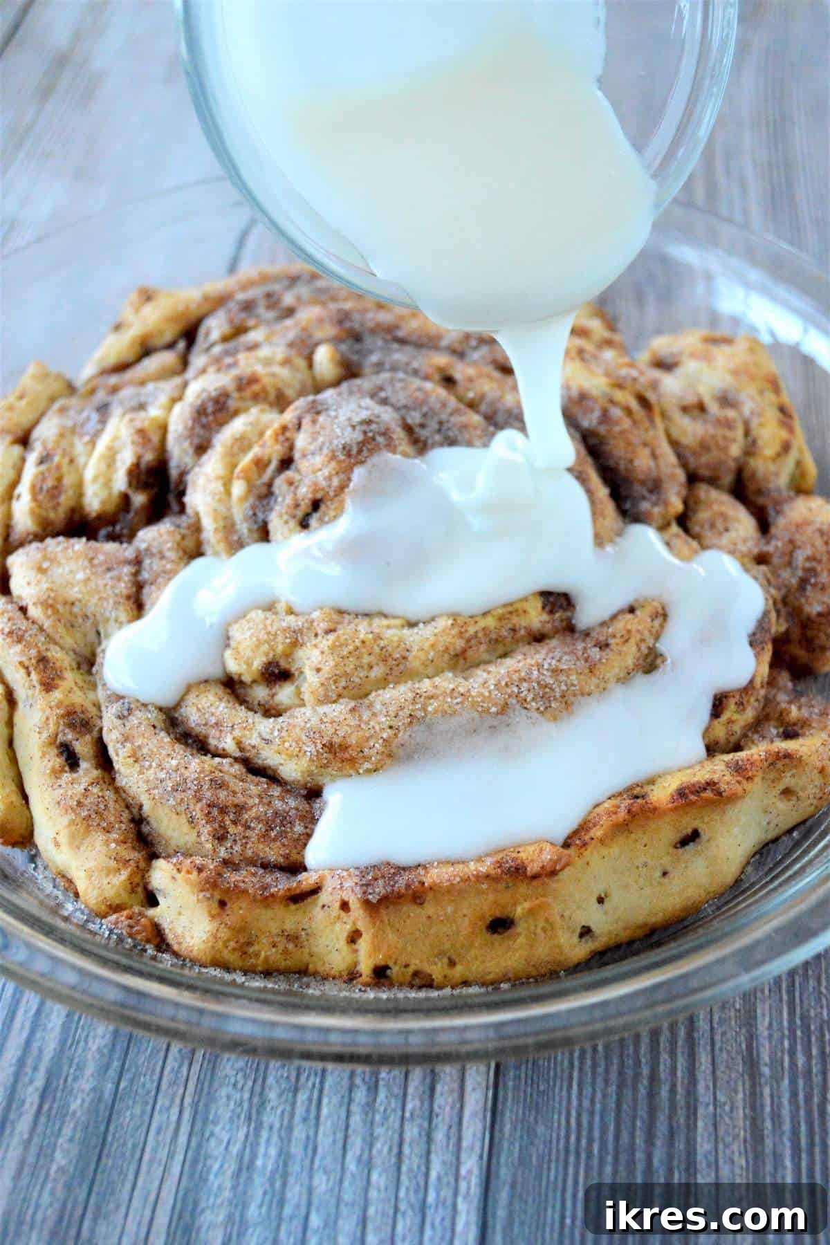 More cream cheese icing being poured, ensuring full coverage of the giant fluffy cinnamon roll.