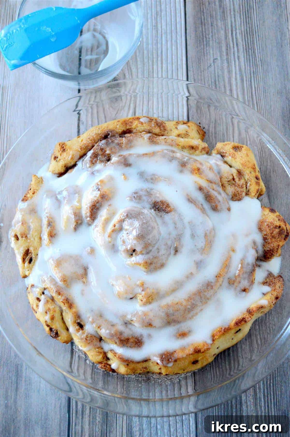 A giant fluffy cinnamon roll completely covered in cream cheese icing, with a spatula on the side for spreading.