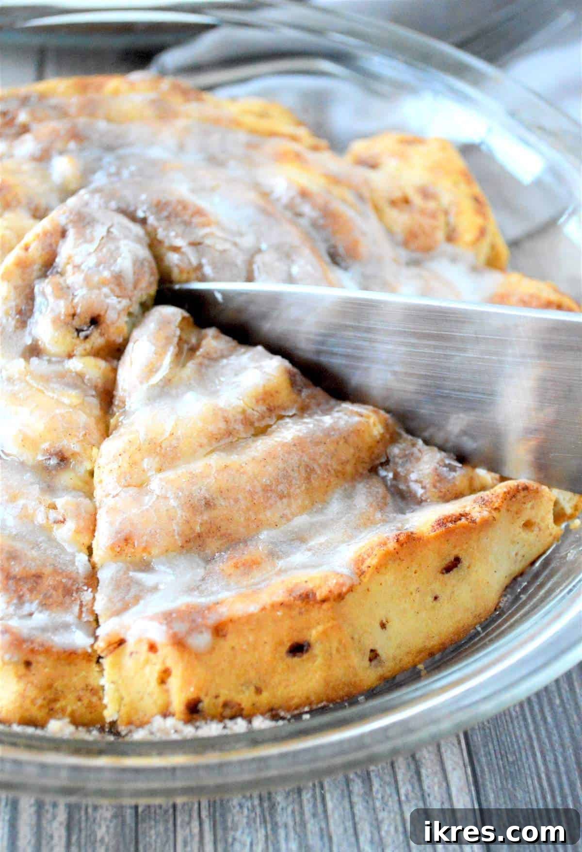 A knife slicing into the center of a giant fluffy cinnamon roll, revealing its soft, warm interior and rich glaze.