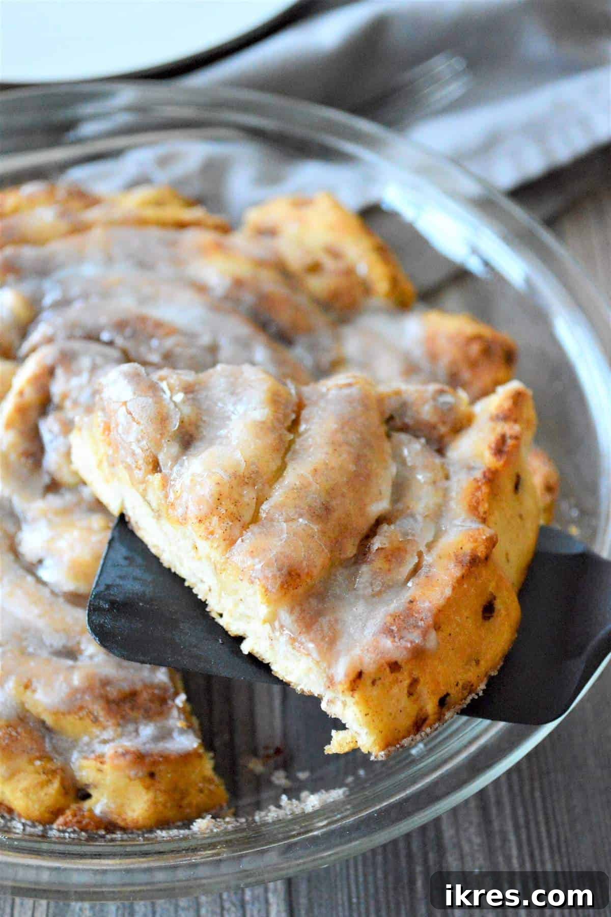 A hand holding a slice of a giant fluffy cinnamon roll over a baking dish, with the remaining large cinnamon roll visible.