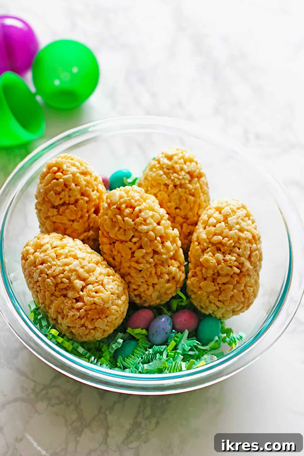 Egg-Cellent Crispy Easter Bites 2 Vibrant Easter eggs made from Rice Krispie Treats, nestled in a clear bowl with festive green Easter grass and colorful candies, showcasing the hidden M&M surprise.