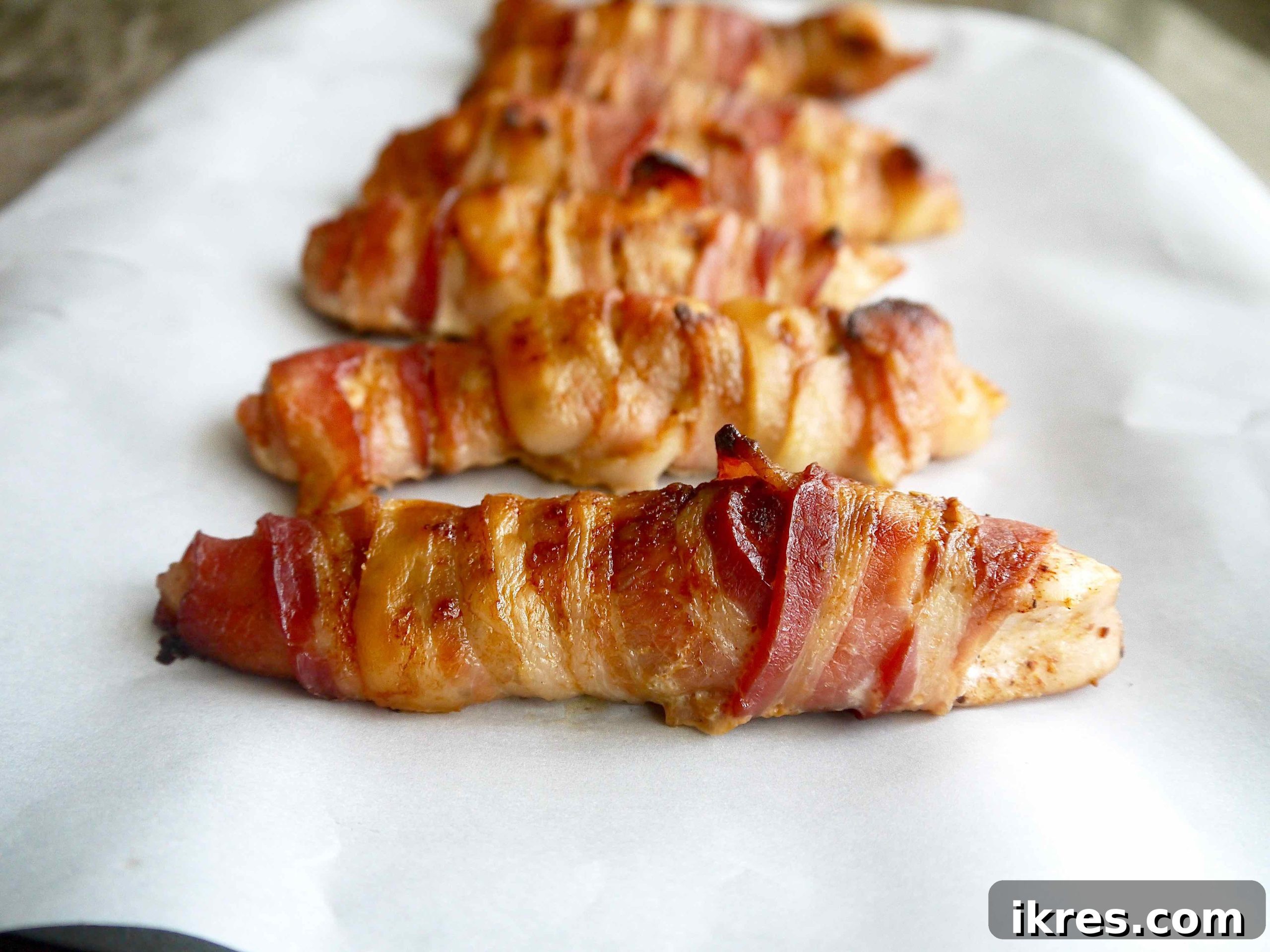 Guilt Free Paleo Keto Bacon Wrapped Chicken Tenders 3 Chicken tenders meticulously wrapped in bacon and arranged on a baking sheet, ready for the oven.