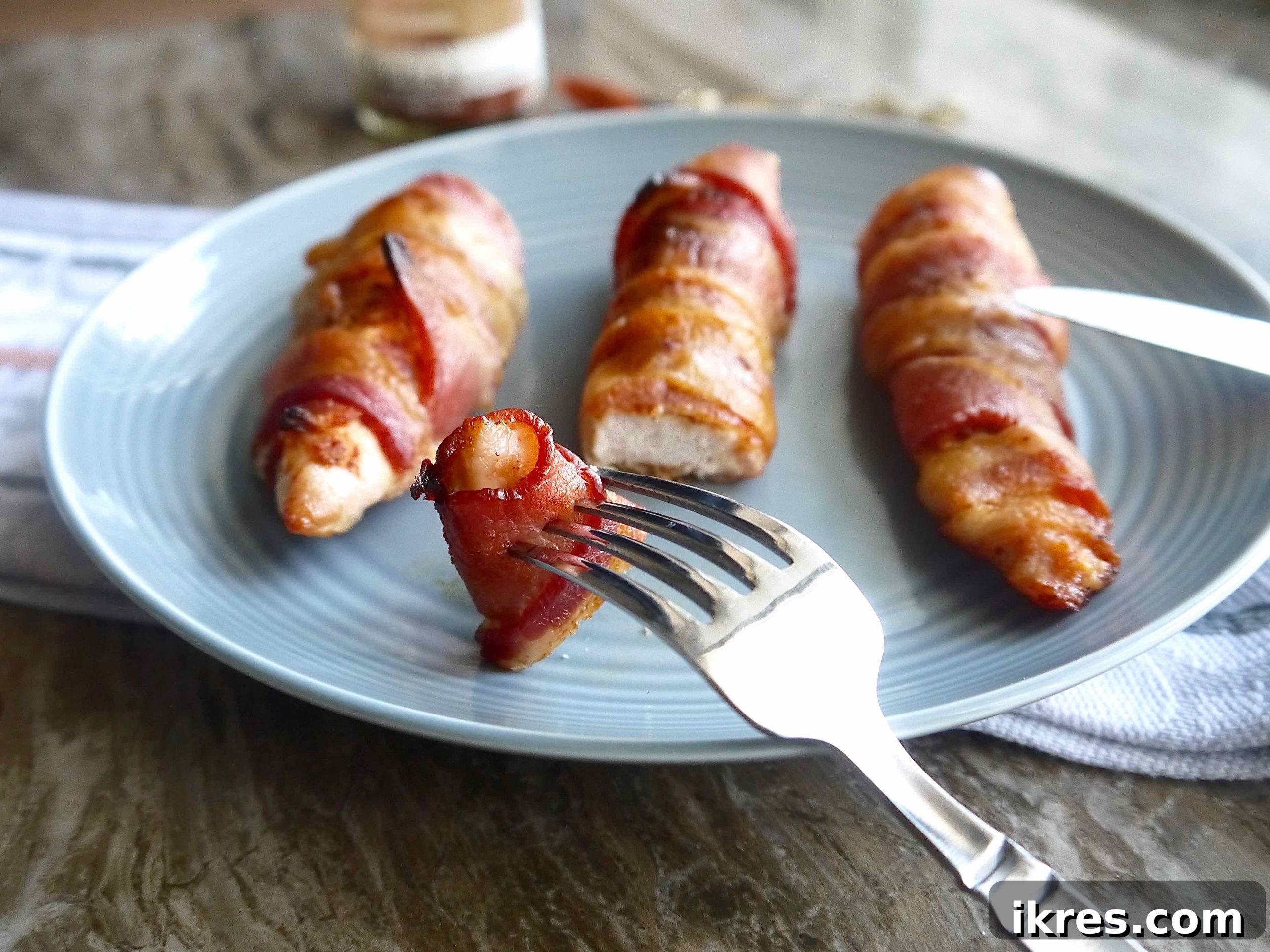 Guilt Free Paleo Keto Bacon Wrapped Chicken Tenders 8 Keto bacon wrapped chicken tenders served on a white plate with a fork and knife, ready to eat.