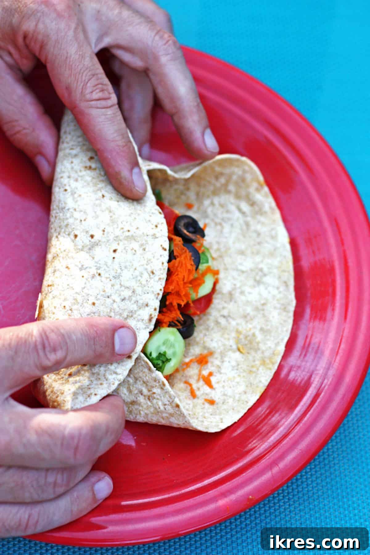 Vibrant Hummus Veggie Wraps 3 Hands carefully rolling up a veggie hummus wrap on a red plate, demonstrating the folding technique.