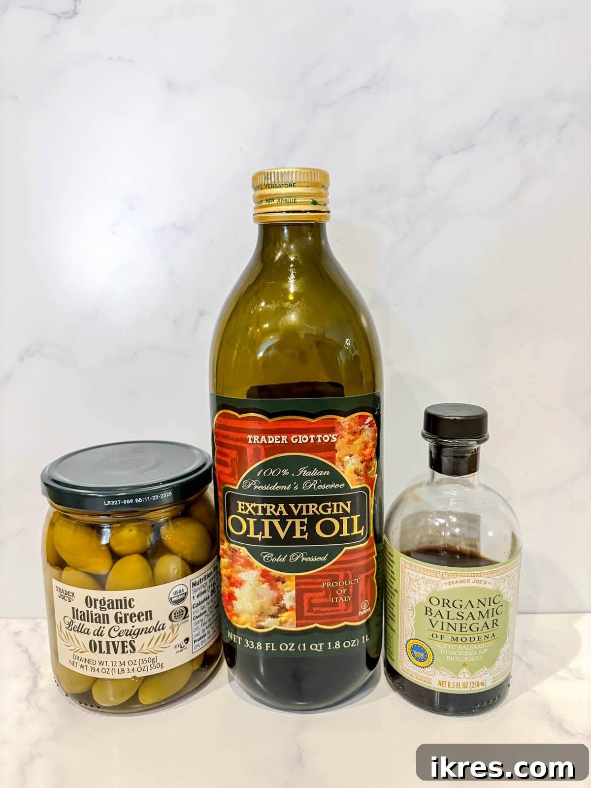 Warm Green Olives Bathed in Balsamic and Olive Oil 3 A carefully arranged selection of essential ingredients for warm green olives, including a jar of whole green olives, bottles of dark balsamic vinegar and vibrant extra virgin olive oil, and fresh oregano sprigs, displayed on a rustic wooden surface.