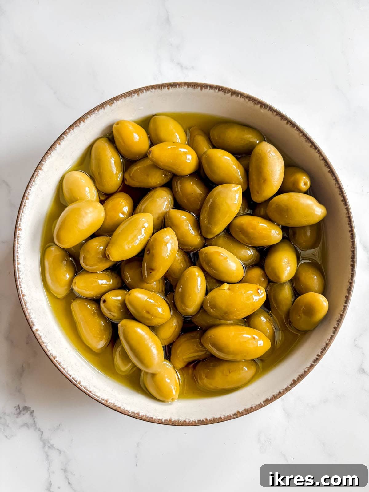 Warm Green Olives Bathed in Balsamic and Olive Oil 4 Drained whole green olives placed in a ceramic oven-safe dish, generously topped with fresh olive oil and balsamic vinegar, ready for baking.